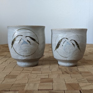 May include: Two white ceramic teacups with a light blue and brown design. The cups have a rounded shape and a small foot.