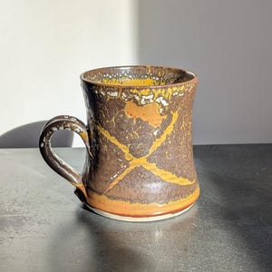 May include: A handmade ceramic mug with a brown and yellow cross design. The mug has a curved handle and a textured surface. The interior and rim are yellow, and the base is orange. The mug is suitable for hot or cold beverages.