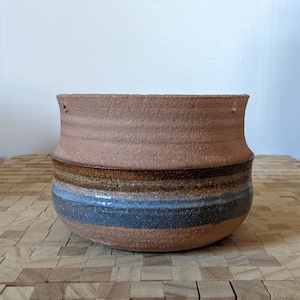 May include: A brown ceramic planter with blue and brown stripes. The planter has a rough, textured surface.