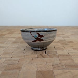 May include: A small, hand-painted ceramic bowl with a gray and white exterior. The bowl features a dark brown rim and a decorative design of brown and teal shapes. The bowl is set on a wooden surface.