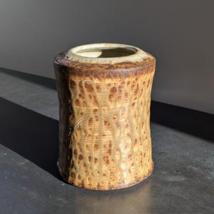 May include: A cylindrical ceramic vase with a textured, dripped glaze in shades of brown and tan. The vase has a wide opening and a decorative, incised pattern near the base. The interior is a lighter tan color.