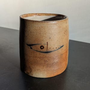 May include: A cylindrical, handmade ceramic mug with a tan and brown speckled finish. A stylized blue line drawing of a fish is on the front. The mug has a slightly irregular shape and a smooth, unglazed interior.