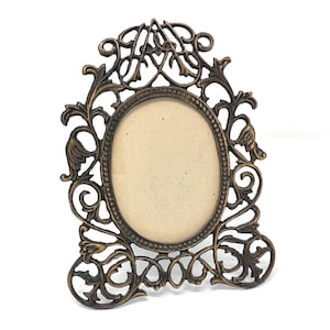 May include: Ornate bronze-colored picture frame with an oval opening. The frame features intricate scrollwork and decorative flourishes around the edges. The frame is designed to stand upright on a flat surface.