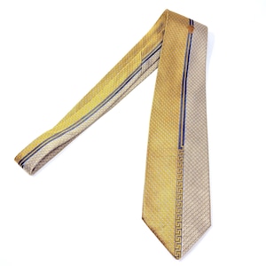 Pode incluir: Uma gravata dourada com um padrão geométrico e uma faixa azul. A gravata tem uma superfície texturizada e uma borda decorativa com um desenho de chave grega. A gravata é exibida em um fundo branco.