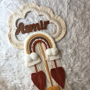 Puede incluir: Un tapiz de macramé de pared con un diseño de arcoíris y un letrero de nombre de madera que dice "Asmir". El arcoíris está hecho de hilo beige, marrón, naranja y amarillo. El tapiz tiene pompones blancos y hojas de macramé marrones y amarillas.