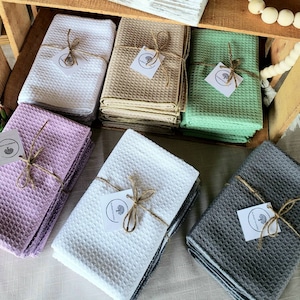May include: Set of six waffle weave kitchen towels in various colors, including white, beige, green, purple, and gray. Each towel is tied with twine and a small tag with the text "Handmade with Love".