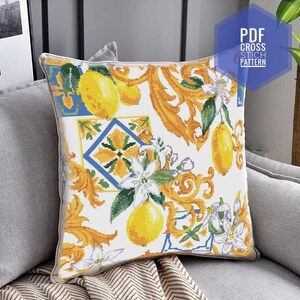 May include: White pillow with a cross stitch pattern of lemons, flowers, and ornate designs in yellow, green, blue, and white.  The pillow is on a gray couch with a brown and white throw blanket.