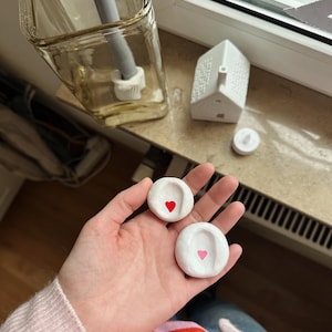 Könnte beinhalten: Zwei kleine, weiße Keramikschalen, jede mit einem Herz in der Mitte bemalt, eines rot und eines rosa. Ein klares Glasgefäß mit einer grauen Kerze und ein weißes Keramikhaus sind ebenfalls zu sehen.