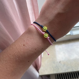 Könnte beinhalten: Zwei Freundschaftsarmbänder mit gelben Smiley-Anhängern. Ein Armband ist pink und das andere ist dunkelblau.