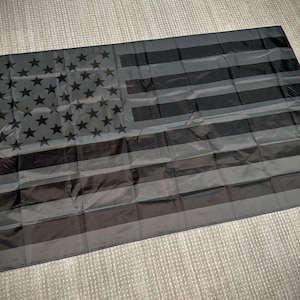 May include: A black and gray American flag with a distressed look. The flag is made of a lightweight material and is perfect for indoor or outdoor use.