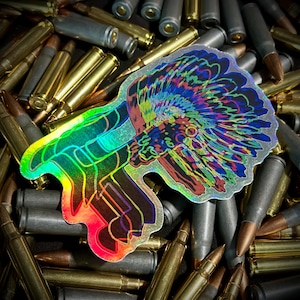 Puede incluir: Una pegatina holográfica con un diseño de tocado nativo americano en colores vibrantes. La pegatina está rodeada de un montón de munición, creando un contraste entre el arte colorido y las balas metálicas.