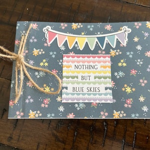 May include: A small, blue notebook with a floral pattern. The notebook has a string tied around it and a sticker that says "Nothing but blue skies" with a rainbow design.