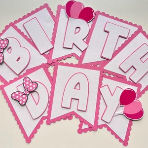 May include: Pink and white birthday banner with the word "Birthday" in white letters on pink flags. Each flag has a scalloped edge. The flags are decorated with pink bows and balloons.