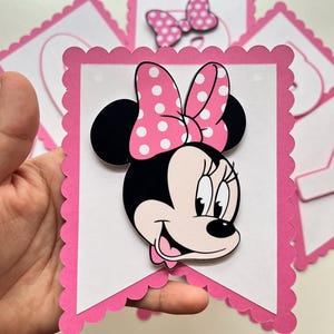 May include: Pink and white banner with a scalloped edge featuring a cartoon image of Minnie Mouse with a pink bow and white polka dots.