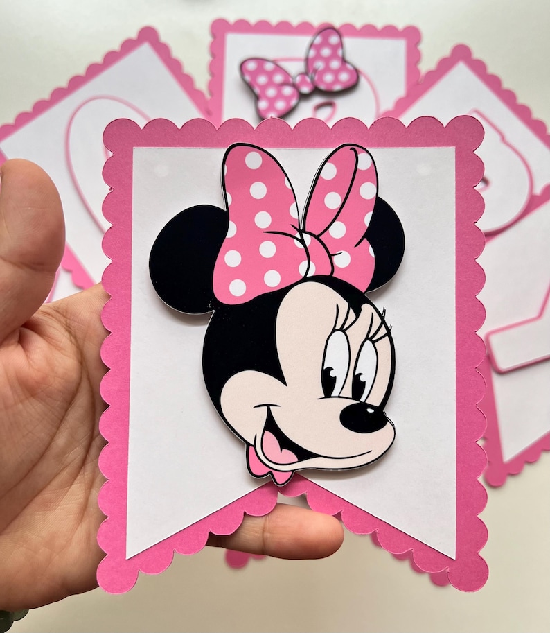 May include: Pink and white banner with a scalloped edge featuring a cartoon image of Minnie Mouse with a pink bow and white polka dots.