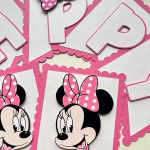 May include: Pink and white banner with a scalloped edge. The banner features a Minnie Mouse character with a pink bow and white polka dots. The banner also has the letter "P" in white with a pink outline.