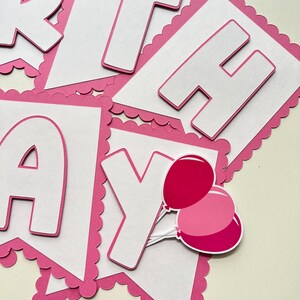 May include: Pink and white paper banner with the word "Birthday" in white letters with a pink outline. The banner has a scalloped edge and includes two pink balloons.