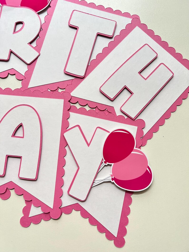 May include: Pink and white paper banner with the word "Birthday" in white letters with a pink outline. The banner has a scalloped edge and includes two pink balloons.