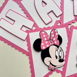 May include: Pink and white banner with scalloped edges featuring the letters "Happy" and a Minnie Mouse character with a red bow and white polka dots.