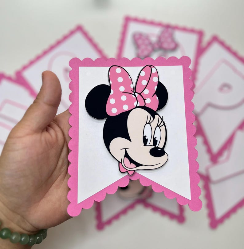 May include: Pink and white banner featuring a Minnie Mouse character. The banner is made from cardstock and has a scalloped edge. The Minnie Mouse character is wearing a pink bow with white polka dots.