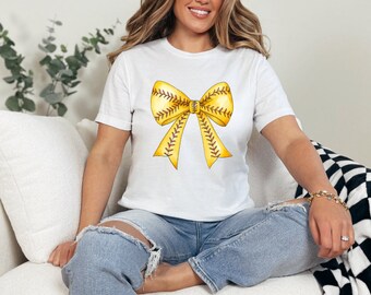 May include: A white short-sleeved t-shirt featuring a large, centered graphic of a yellow bow. The bow is designed with the texture and red stitching pattern of a baseball, creating a distinct sports-themed visual element on the front.