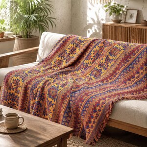 May include: A woven throw blanket featuring a repeating pattern of geometric shapes and floral motifs in vibrant shades of yellow, deep red, and blue. The blanket has a textured surface with a fringed edge, draped over a light-coloured sofa.