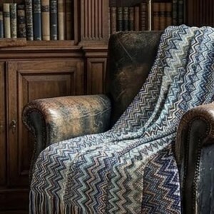 May include: A woven throw blanket featuring a multi-coloured chevron pattern in shades of blue, grey, and cream. The blanket has a textured knit construction and is finished with long, straight fringe along the bottom edge, draped over a chair.