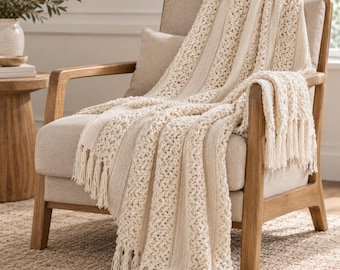 May include: A cream-coloured knit throw blanket featuring alternating vertical panels of open-work crochet and solid ribbed texture, finished with long fringe tassels along the bottom edge, draped over a wooden armchair.