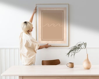 May include: A person hangs a framed print with a beige background and a white line drawing of waves on a white wall. The print is in a gold frame. A wooden table with a vase and a mug is in the foreground.