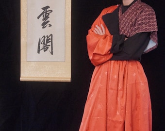 Könnte beinhalten: Eine Person trägt ein rotes und schwarzes Outfit mit einem langen, fließenden Rock. Der Rock hat ein Blumenmuster und ist in der Taille gebunden. Die Person steht vor einer Schriftrolle mit chinesischen Schriftzeichen.