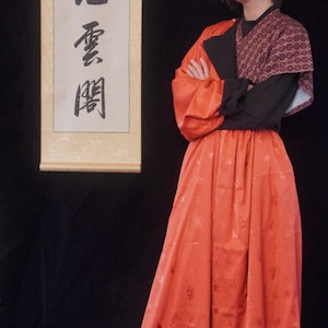 Puede incluir: Una persona que lleva un atuendo rojo y negro con una falda larga y fluida. La falda tiene un estampado floral y está ceñida a la cintura. La persona está de pie frente a un rollo con caracteres chinos.