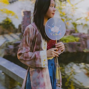 Puede incluir: Una joven con una larga túnica de estampado rosa y marrón sostiene un abanico redondo blanco con un diseño floral. El abanico tiene un mango largo, delgado y de madera con un borla.