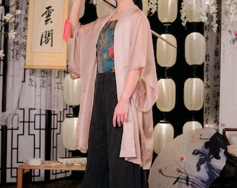 May include: A woman in a pink kimono holds a sword over her shoulder. She is wearing black trousers and a blue patterned top. The background features white flowers and lanterns.