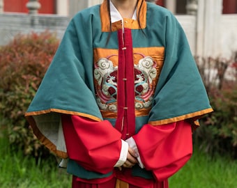 Könnte beinhalten: Eine junge Frau trägt eine traditionelle chinesische Robe mit einem tealgrünen Oberteil, roten Ärmeln und einem roten Rock. Die Robe hat goldene Verzierungen und aufwendige Stickereien mit einem Wolken- und Phönix-Design. Die Frau lächelt und steht vor einem roten und weißen Gebäude.