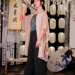 Puede incluir: Una mujer con un kimono rosa sostiene una espada sobre su hombro. Lleva pantalones negros y una blusa con estampado azul. El fondo presenta flores blancas y linternas.