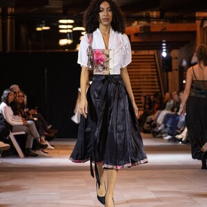 Puede incluir: Una modelo camina por la pasarela con una blusa blanca con estampado floral y una falda negra. La falda tiene un ribete decorativo y un lazo en la cintura.