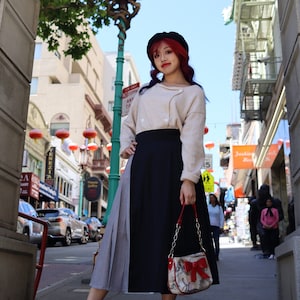 Puede incluir: Una mujer con el pelo rojo y un gorro negro está usando un suéter beige, una falda negra y gris y un bolso de mano rojo y blanco. Está de pie frente a un arco chino con linternas rojas colgando del techo.