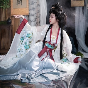 Puede incluir: Una mujer con un vestido tradicional chino blanco y rojo con un diseño de serpiente en la manga. Está sentada en una superficie plateada con un rollo detrás de ella.