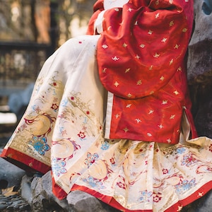 Puede incluir: Una mujer vestida con un vestido tradicional chino con la parte superior roja y la inferior blanca con bordados florales dorados y azules. El vestido tiene una falda larga y fluida y una parte superior ajustada con un cuello alto. La mujer sostiene un abanico en su mano.