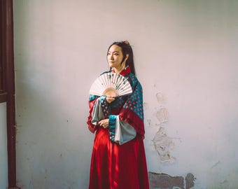 May include: A long-sleeved garment featuring a deep red floor-length skirt and a teal bodice with a repeating pattern of small red shapes. The sleeves are wide and light gray, and the garment is held with a white folding fan with a painted design.
