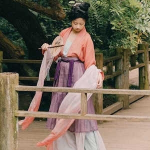 Puede incluir: Una mujer con un vestido tradicional chino rosa y morado se encuentra de pie en un puente de madera en un entorno de jardín. Sostiene un abanico en su mano derecha y una larga bufanda que fluye en su mano izquierda.