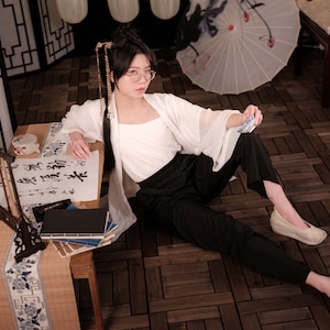 Puede incluir: Una mujer con un top blanco y pantalones negros se sienta en un suelo de madera. Ella sostiene un trozo de papel en su mano. En primer plano, una mesa con una superficie de madera y un camino de mesa de bambú. La mesa tiene un juego de caligrafía y un libro.