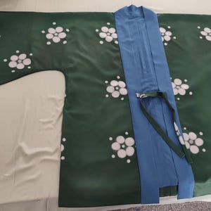 Puede incluir: Bata estilo kimono verde con estampados florales blancos. La bata presenta una solapa azul y una corbata a juego. La prenda está hecha de un material sedoso y está tendida sobre una superficie clara.