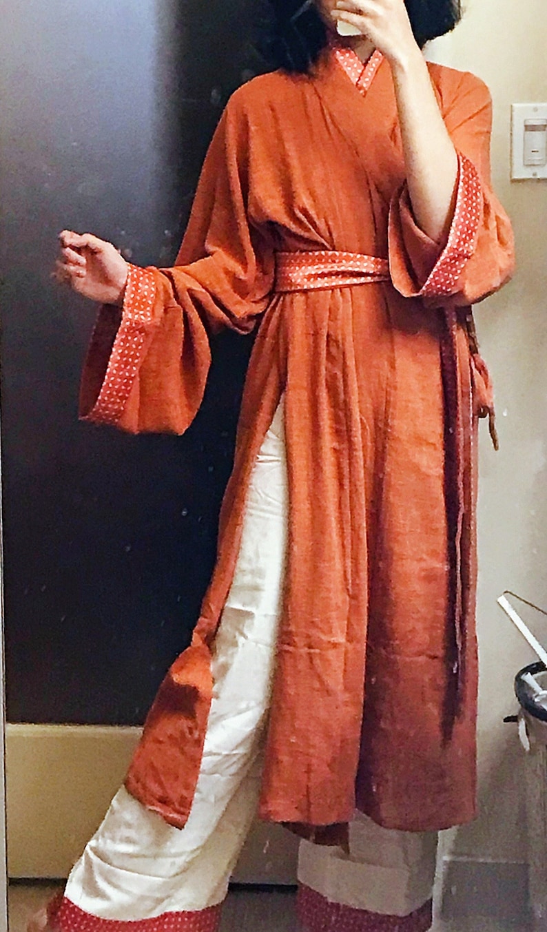 May include: A long, rust-coloured robe with a white sash and white trousers. The robe has a V-neck and long sleeves with red and white polka dot trim. The trousers have a red and white polka dot trim at the bottom.