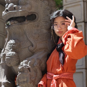 May include: A young person wearing a long, orange robe with a white patterned sash and a pair of gold-rimmed glasses. The person is standing next to a large, grey stone lion statue.