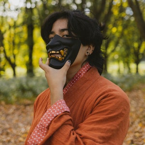 May include: A person wearing a black and gold oni mask with a red and white patterned robe. The person is standing in a forest with autumn foliage.