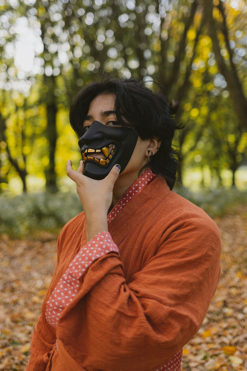 May include: A person wearing a black and gold oni mask with a red and white patterned robe. The person is standing in a forest with autumn foliage.