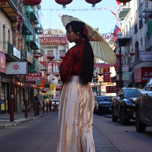 May include: A woman wearing a red and gold traditional Chinese dress and a white parasol walks down a street lined with red lanterns.