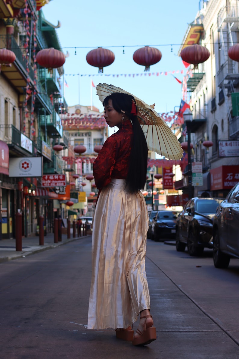 May include: A woman wearing a red and gold traditional Chinese dress and a white parasol walks down a street lined with red lanterns.