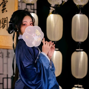 May include: A woman in a blue robe holds a white fan with a rabbit design in front of her face. The fan has a gold handle. The background is a wall with white lanterns.